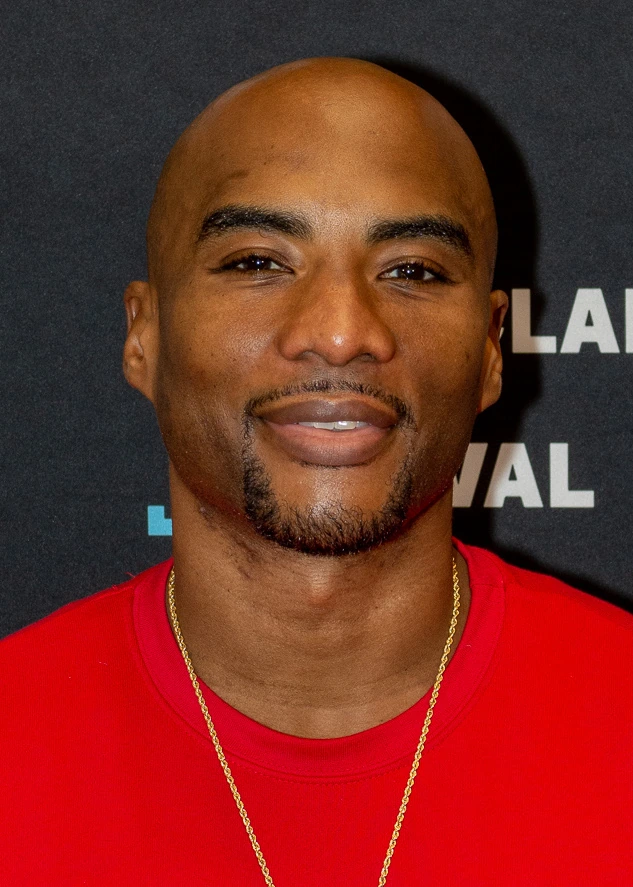 Charlamagne Tha God at a public event, wearing a red shirt and gold chain, posed against a dark backdrop